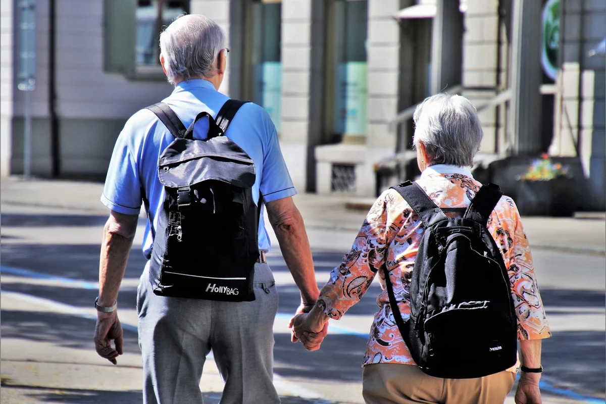 Invecchiamento attivo, due anziani per mano camminano con zaino da viaggio