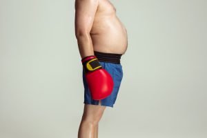 Obesità: un uomo di profilo con capelli rossi e pancia scoperta che sporge in vista indossa guantoni rossi da box e pantaloncini blue