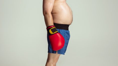 Obesità: un uomo di profilo con capelli rossi e pancia scoperta che sporge in vista indossa guantoni rossi da box e pantaloncini blue