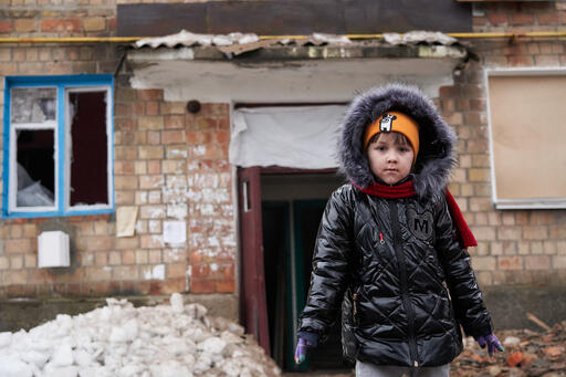 giornata mondiale infanzia, una bimba con alle spalle la casa distrutta dalla guerra in Ucraina